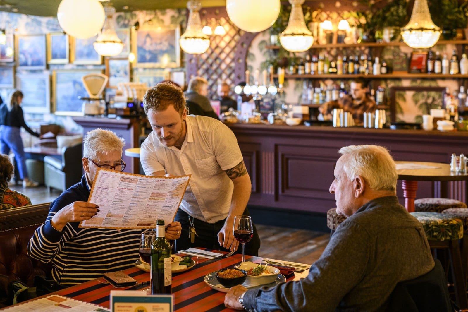 A waiter assists an elderly woman holding a menu at a restaurant table, as an elderly man sits across from her with food, a coltello, and a glass of red wine. The bar and other diners are visible in the background.