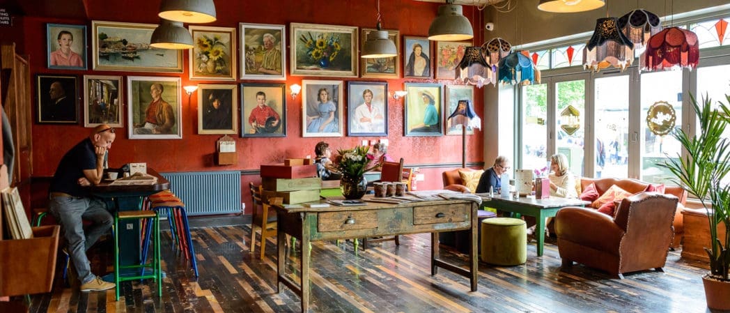 A cozy café in Como with red walls, vintage furniture, and colorful portraits above. People sit at tables and sofas, chatting and working. Pendant lamps and natural light from large windows create a warm, inviting atmosphere.