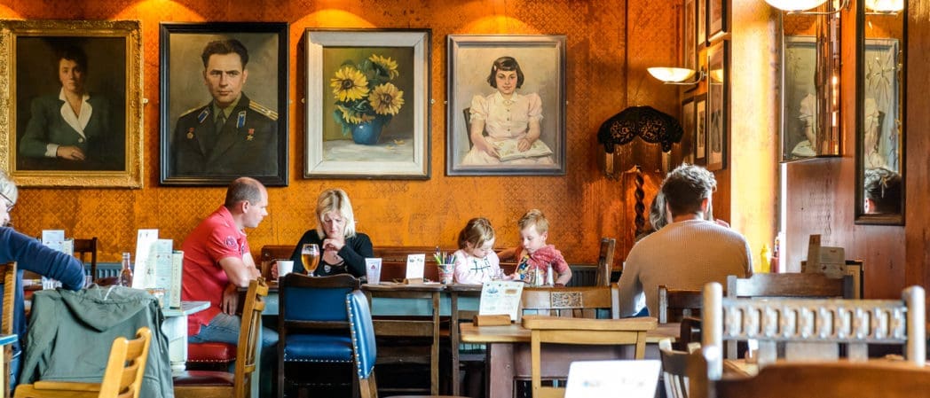 People sit at tables in a cozy Como café with wooden furniture and orange patterned walls decorated with large, framed portraits and paintings. Some diners are eating, drinking, or talking, and two young children are present.