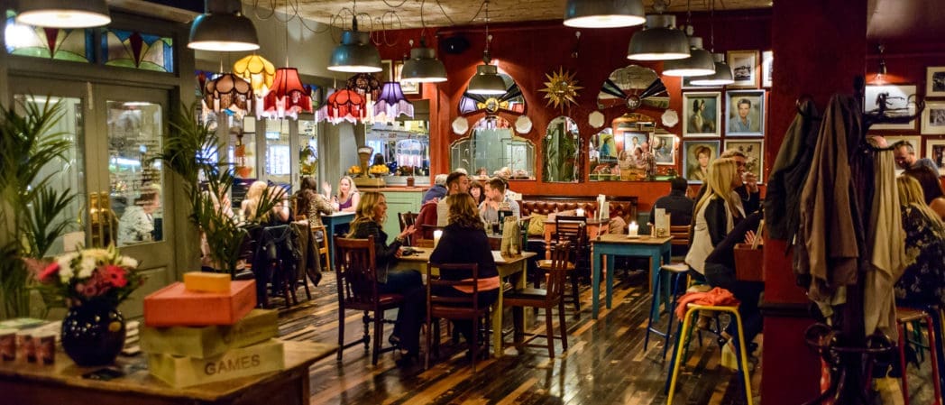 A cozy, eclectic restaurant interior in Como with colorful hanging lamps, wooden floors, and groups of people dining at various tables. Walls are decorated with framed pictures and mirrors, creating a warm, inviting atmosphere.