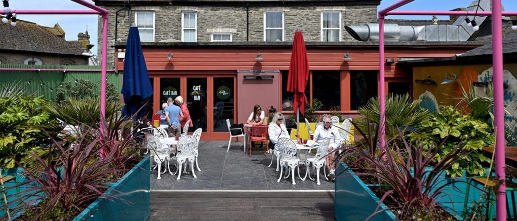 People sit at colorful outdoor tables in front of a café with a red awning and large Concho-style windows. Decorative plants and umbrellas line the seating area, with a two-story brick building in the background.