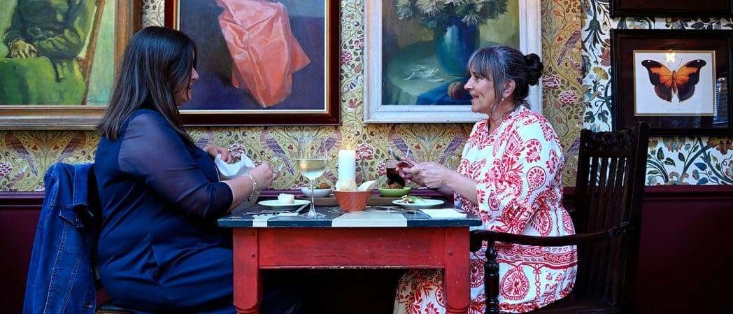 Two women sit across from each other at a small table in a warmly decorated Condado room with framed art on the walls, sharing a meal and conversation by candlelight.