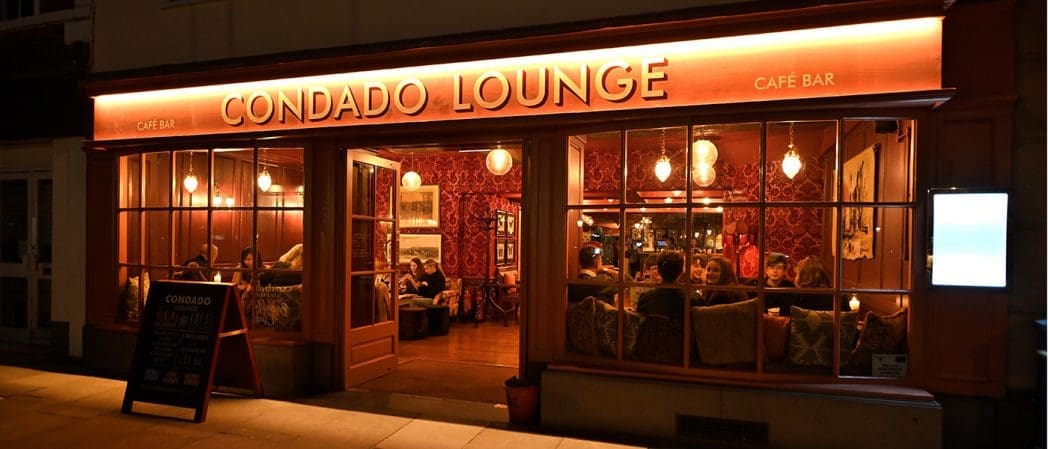 Exterior view of Condado Lounge café bar at night, warmly lit with an orange glow. People are sitting inside on sofas, visible through large windows. A Condado signboard stands outside on the pavement.