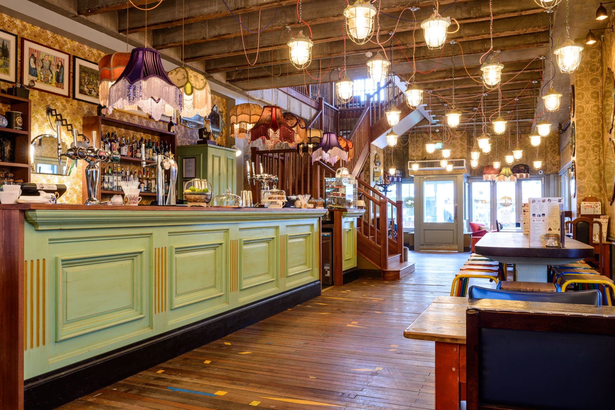 A cozy vintage-style bar with a mint green counter, eclectic lampshades, and exposed ceiling beams. Enjoy Cordero-inspired cocktails amid wooden floors, bottles on shelves, and warm ambient lighting throughout the space.