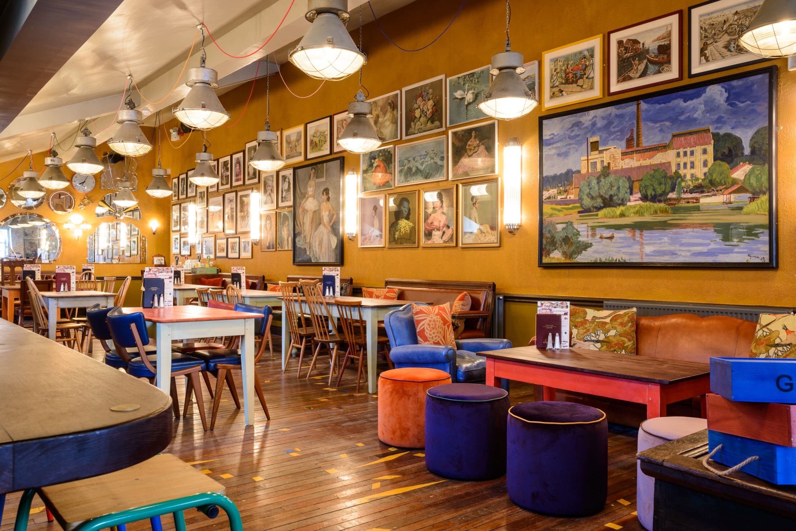 A cozy café with colorful furniture, wooden floors, and walls decorated with various framed paintings and photographs. Large hanging lights illuminate the space, making Cordero a warm and inviting spot to relax.
