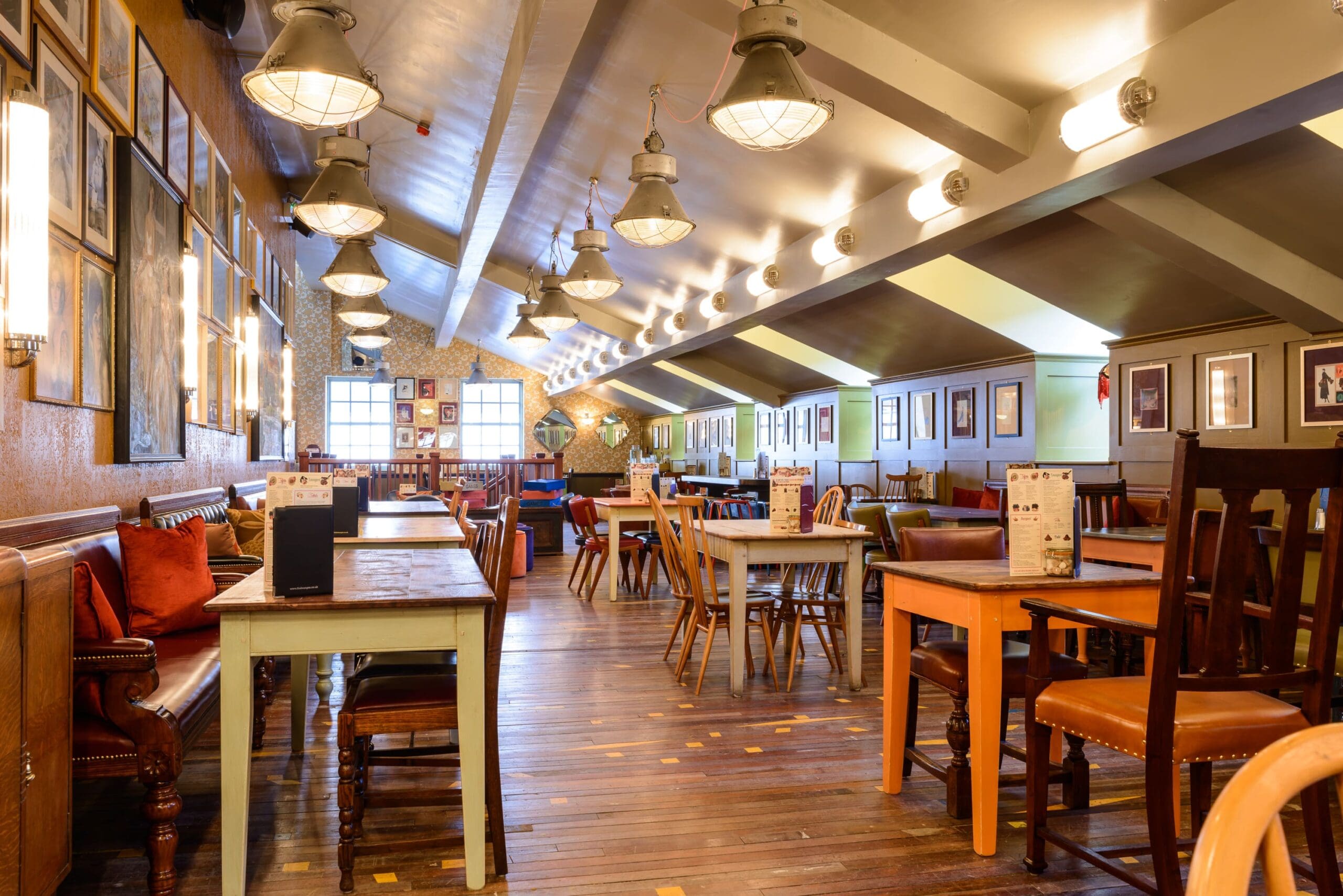 A brightly lit restaurant with wooden floors, assorted wooden tables and chairs, wall art, and pendant ceiling lights. The space appears empty and inviting, with menus featuring specialties like cordero placed on each table.