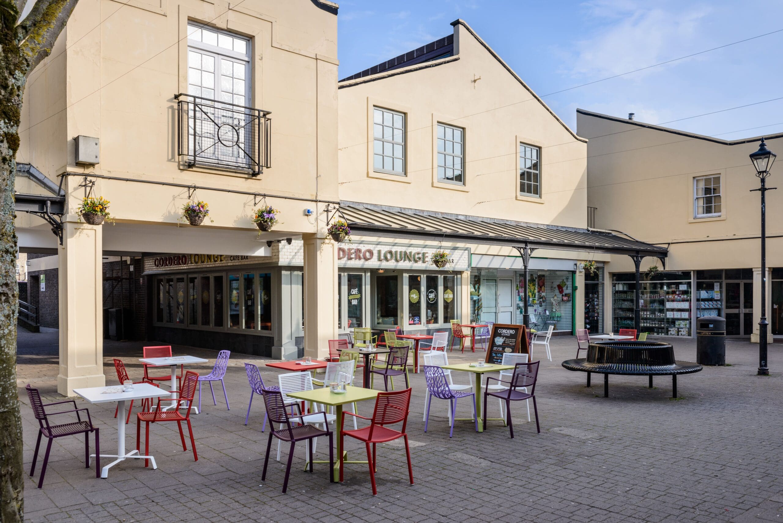 An outdoor plaza with colorful chairs and tables in front of "Dero Lounge" café. Nearby are shops, a round bench with a lamppost, and light-colored Cordero buildings. The well-lit area is empty and inviting.