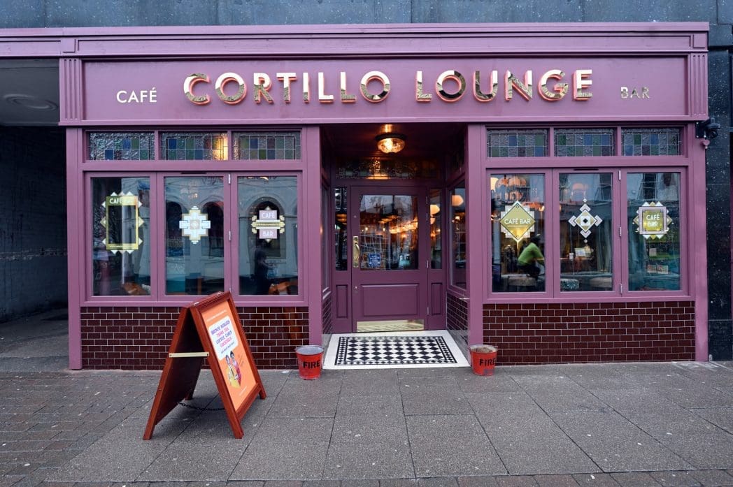 Front view of Cortillo Lounge café bar with its purple and brown exterior, large windows, a decorative tiled entrance, and a sidewalk sign welcoming you in front of the door.