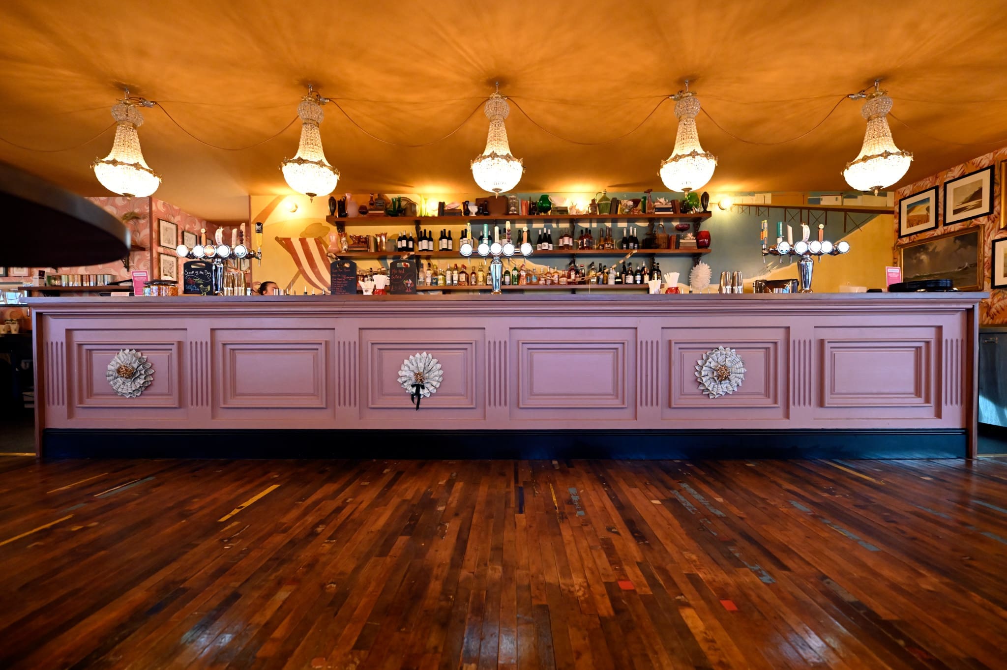 A stylish bar, Costero, features a long pink paneled counter, ornate chandeliers overhead, shelves stocked with bottles and glasses, and a polished wooden floor.