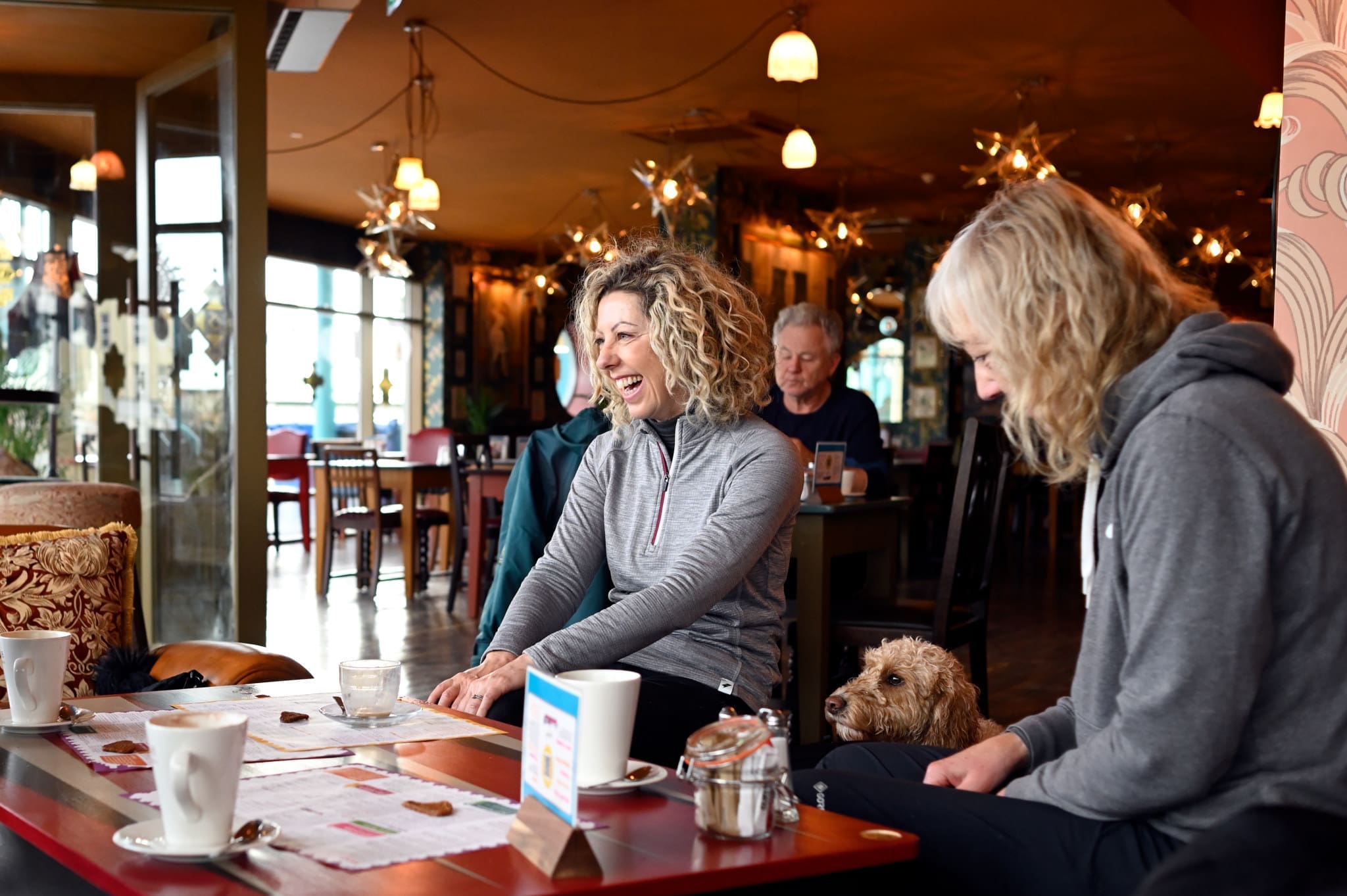 Two women sit at a cozy Costero café table, smiling and chatting. A small brown dog sits beside one of them. Coffee cups and a sugar jar rest on the table, surrounded by warm lighting and a welcoming atmosphere.