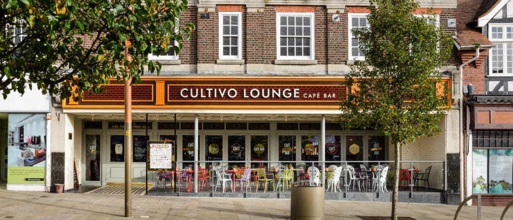 A street view of Cultivo Lounge Café Bar with outdoor seating, colorful chairs and tables, and large windows. The Cultivo sign stands out against the brickwork above, partially hidden by leafy trees in the foreground.