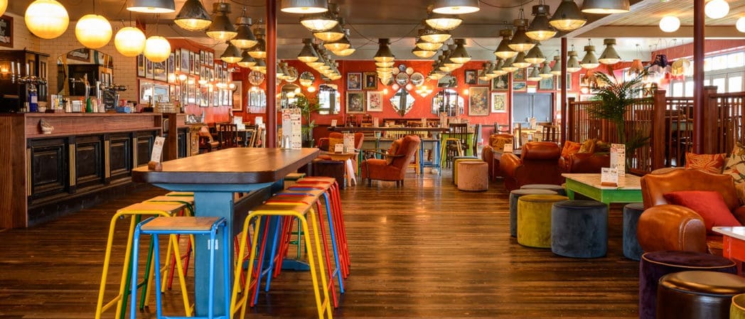 A colorful, cozy bar by Cultivo with wooden floors, vibrant stools, comfy chairs, warm lighting, and eclectic wall art. The space appears inviting, spacious, and stylish, blending modern and retro decor elements.