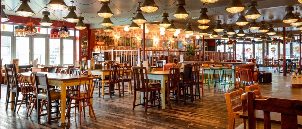 A spacious, well-lit Cultivo restaurant interior with wooden floors, colorful mismatched tables and chairs, vintage-style hanging lights, and framed pictures on the walls. The empty space creates a welcoming, cozy atmosphere.