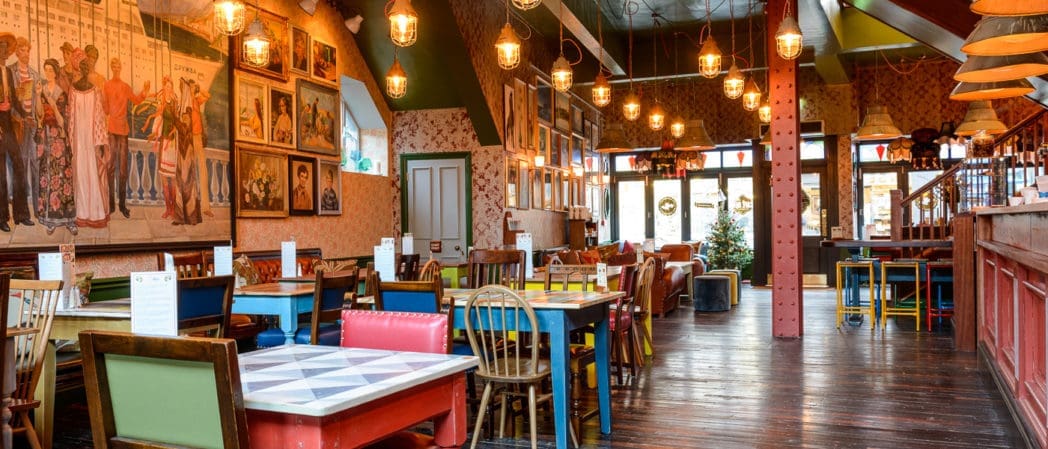Colorful, eclectic restaurant interior with mismatched chairs and tables, warm hanging lights, framed artwork, and eye-catching curio displays. Large windows fill the space with natural light for a cozy and inviting atmosphere.