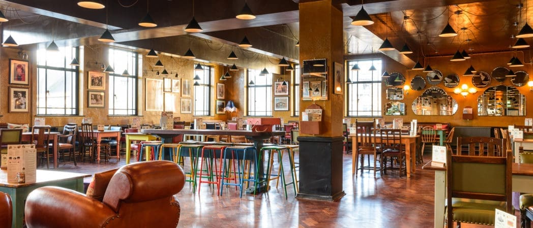 Spacious, well-lit restaurant with wooden floors, eclectic chairs and tables, Curio bar stools, framed pictures on the walls, and large windows letting in natural light.