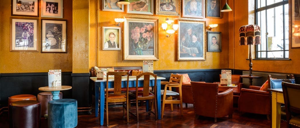 Cozy café with mustard yellow walls, eclectic armchairs and tables, vintage lamps, and various curio pieces among the framed portraits and paintings. Sunlight streams in through a large window, creating a warm, inviting atmosphere.