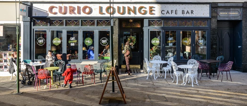 The outdoor seating area of Curio Lounge Café Bar features colorful chairs and tables. People are sitting and talking; a child in a red outfit walks toward the Curio entrance. Large windows and decorative signage add to the lively scene.