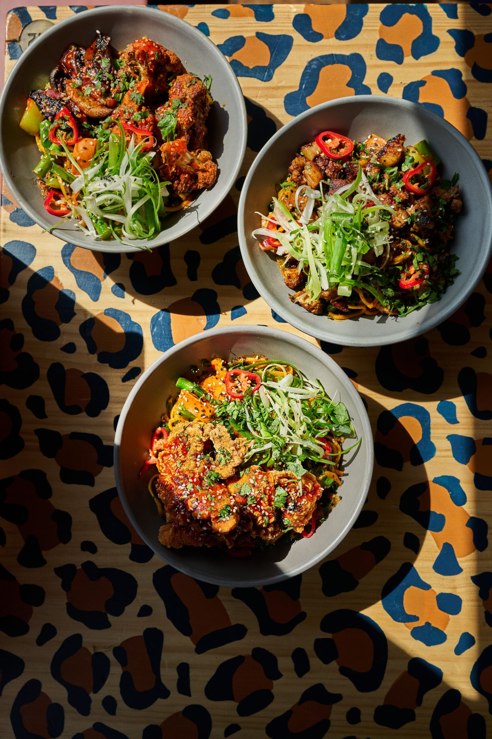 Three bowls of colorful salads topped with fried chicken, sliced red chili, green onions, and sesame seeds, arranged on a table with a bold, blue and orange animal print pattern.
