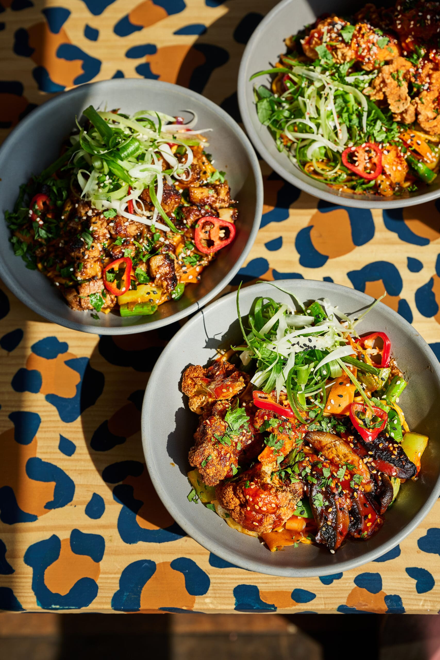 Three bowls of colorful, garnished food, including fried items, sliced red chili, fresh herbs, and greens, are arranged on a spotted animal print-patterned table in a vibrant lounge setting. Bright lighting highlights the fresh ingredients.