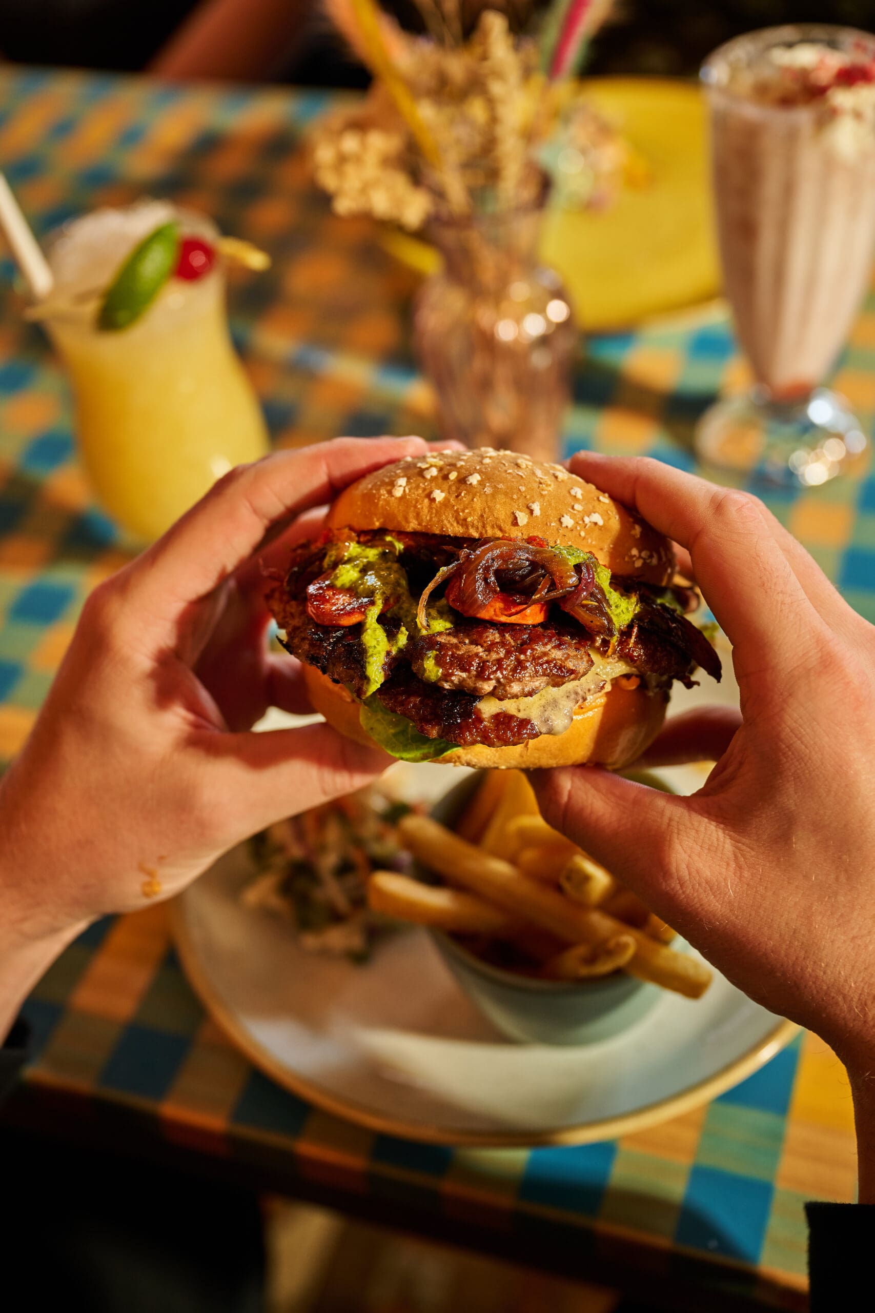 A person holds a burger with lettuce, tomato, and grilled vegetables above a plate of French fries. On the table are a smoothie, a milkshake, and a checkered tablecloth with floral decorations in the background.