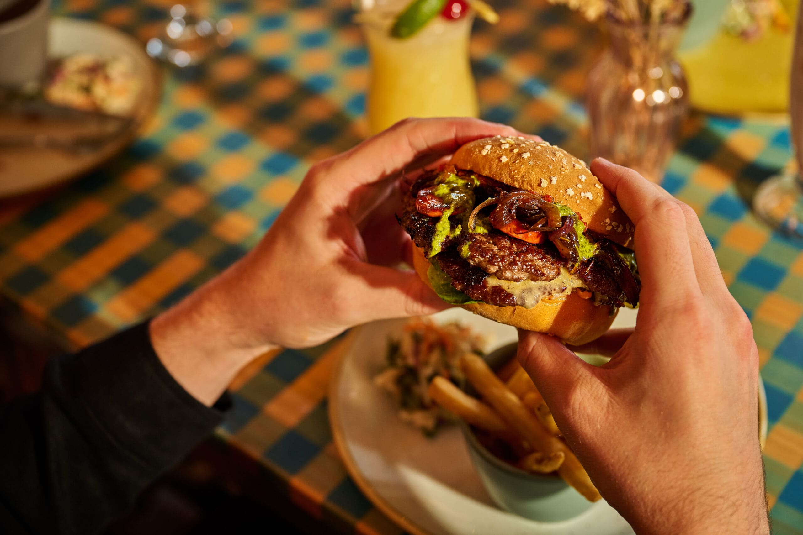A person holding a hamburger with lettuce, tomato, and sauce, above a plate of fries on a checkered tablecloth, with drinks and flowers in the background.