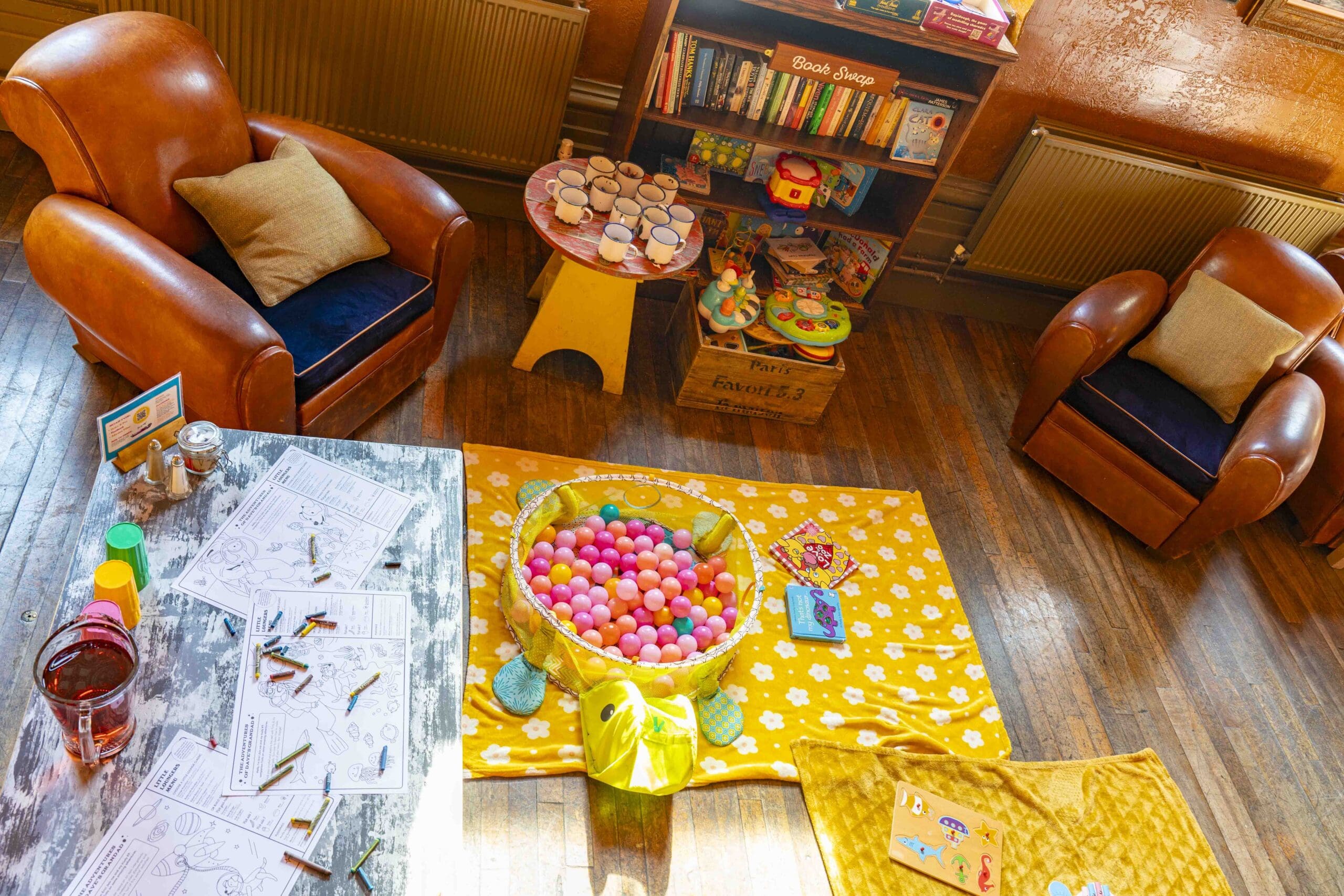 A cozy playroom with two armchairs, a bookshelf, coloring pages and crayons on a table, a basket of colorful balls on a yellow mat, and various toys arranged on wooden flooring.