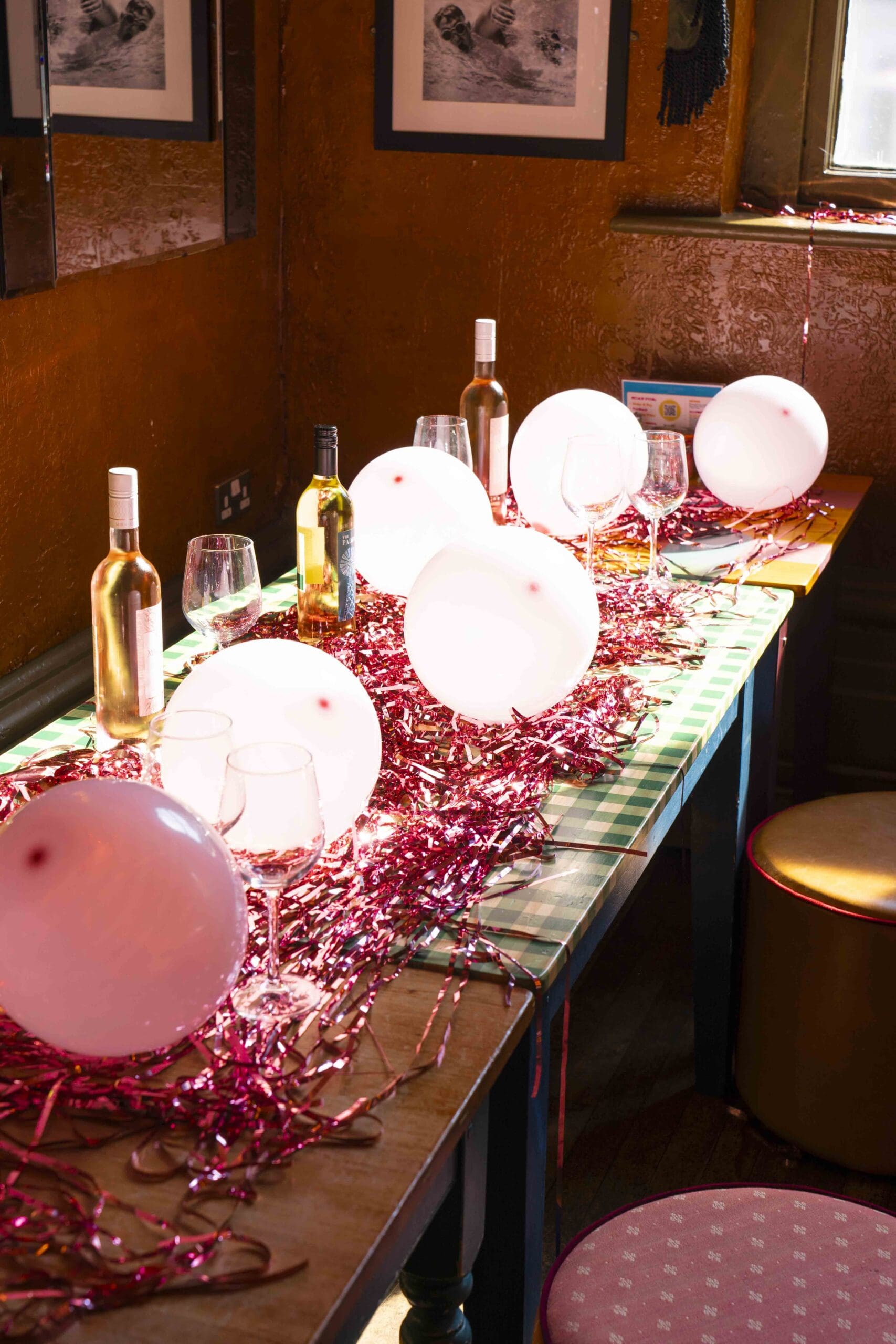 A table decorated with pink balloons, shiny pink streamers, wine bottles, and glasses, set in a warmly lit room with framed pictures on the wall and sunlight streaming in through a window.