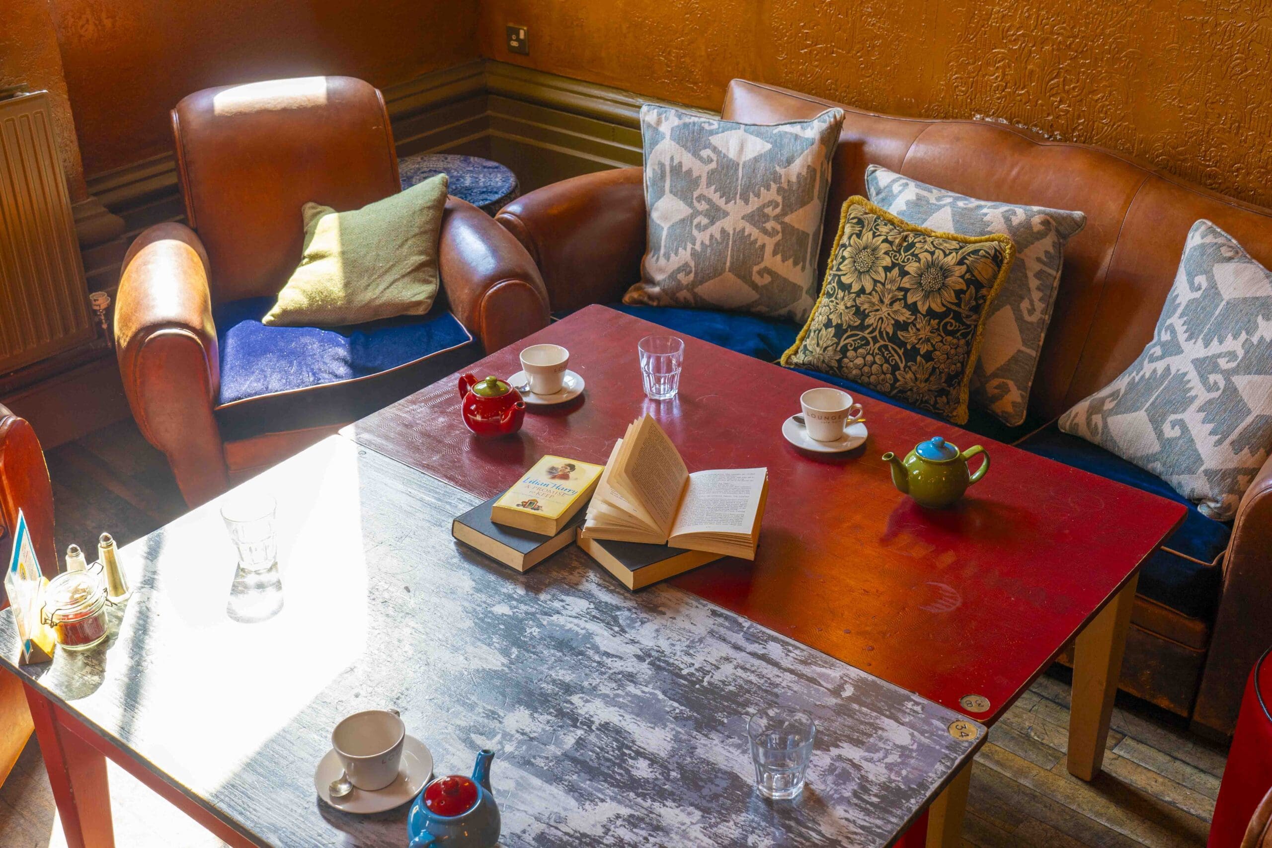 A cozy seating area with leather chairs, a sofa with patterned pillows, a red table holding teapots, cups, glasses, and open books, bathed in warm sunlight from the side.