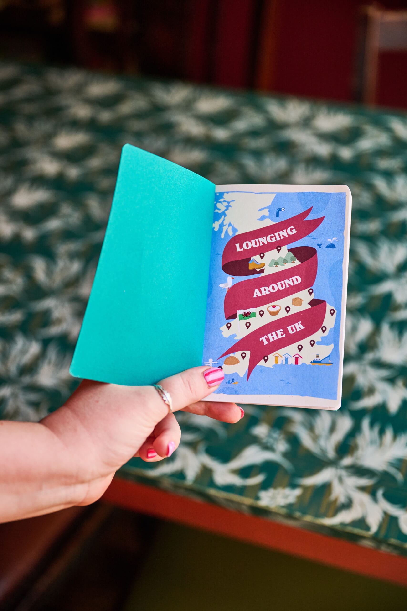 A person with pink nail polish holds an open booklet showing an illustrated page that reads, "Lounging Around the UK" with travel-themed icons on a leafy-patterned table.