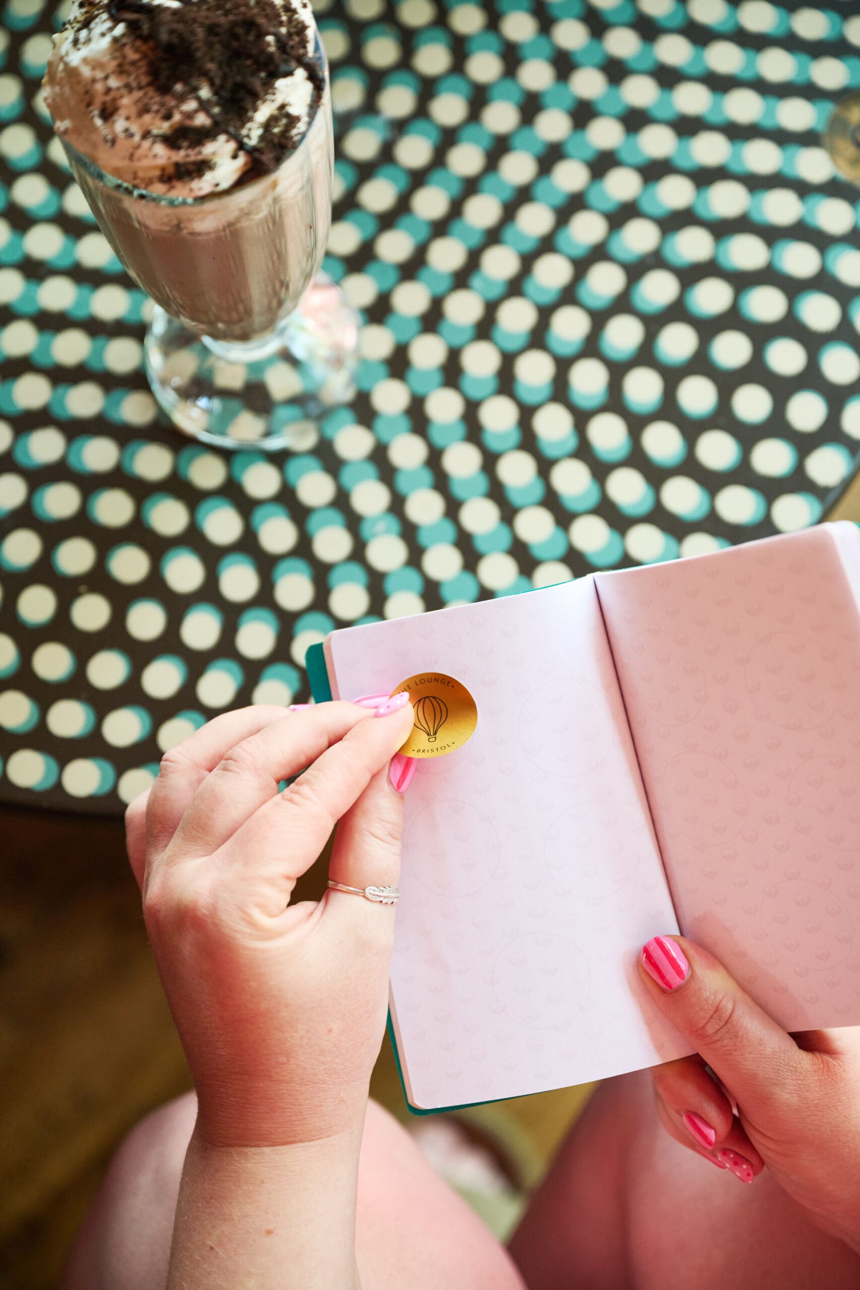 A person with pink nail polish peels a gold sticker in a notebook with faint pink hearts, sitting at a polka-dotted table with a glass of chocolate milkshake topped with whipped cream and chocolate shavings.