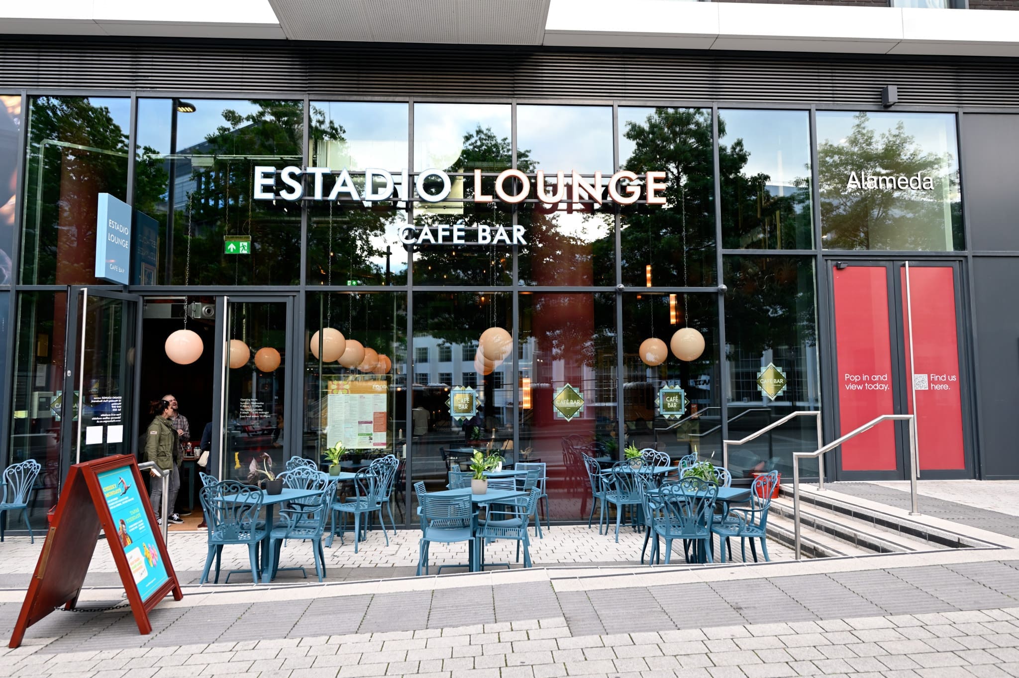Street view of Estadio Lounge Café Bar with glass front, blue outdoor chairs and tables arranged on the pavement, large round lights inside, and an Estadio signboard near the entrance.