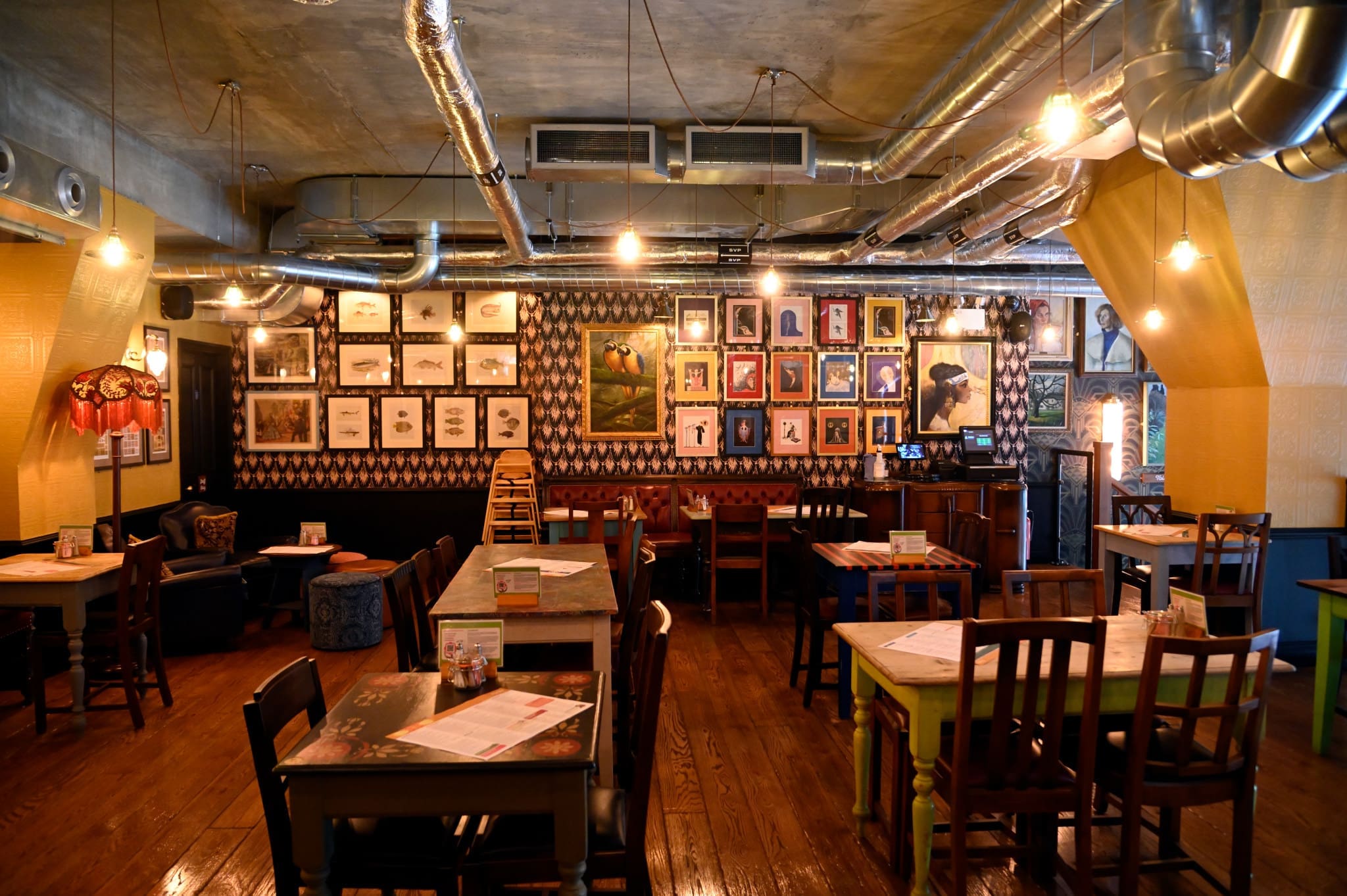 A cozy restaurant interior near Estadio, with wooden floors, mismatched tables and chairs, exposed ductwork, and framed pictures on the walls. Warm lighting creates an inviting atmosphere, and no people are present.