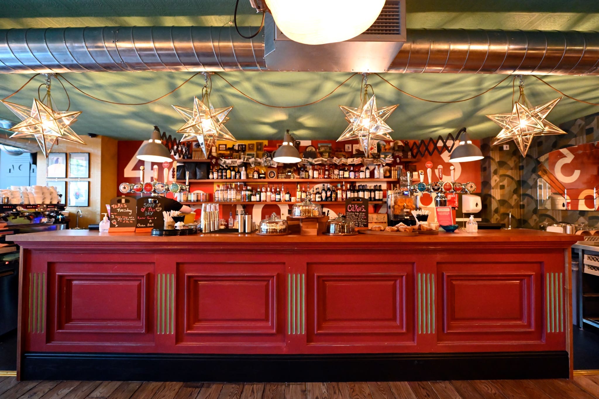 A vibrant café bar near Estadio, featuring a red counter, hanging star-shaped lights, and shelves stocked with bottles and various items. Metal air ducts run along the green ceiling above the warm, inviting setting.