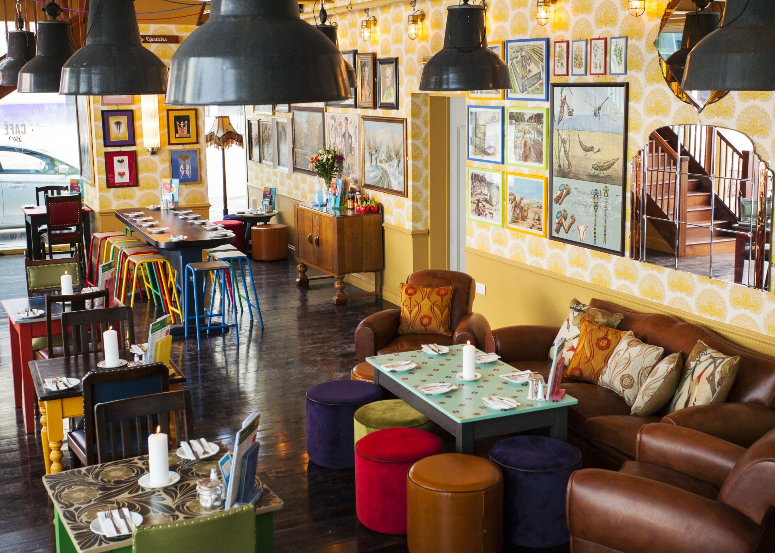 A colorful, eclectic café interior with mismatched chairs, cozy sofas, vibrant tables, and unique Falco-inspired wall art. Hanging lights and candles add to the warm, inviting atmosphere with creative table settings throughout the space.