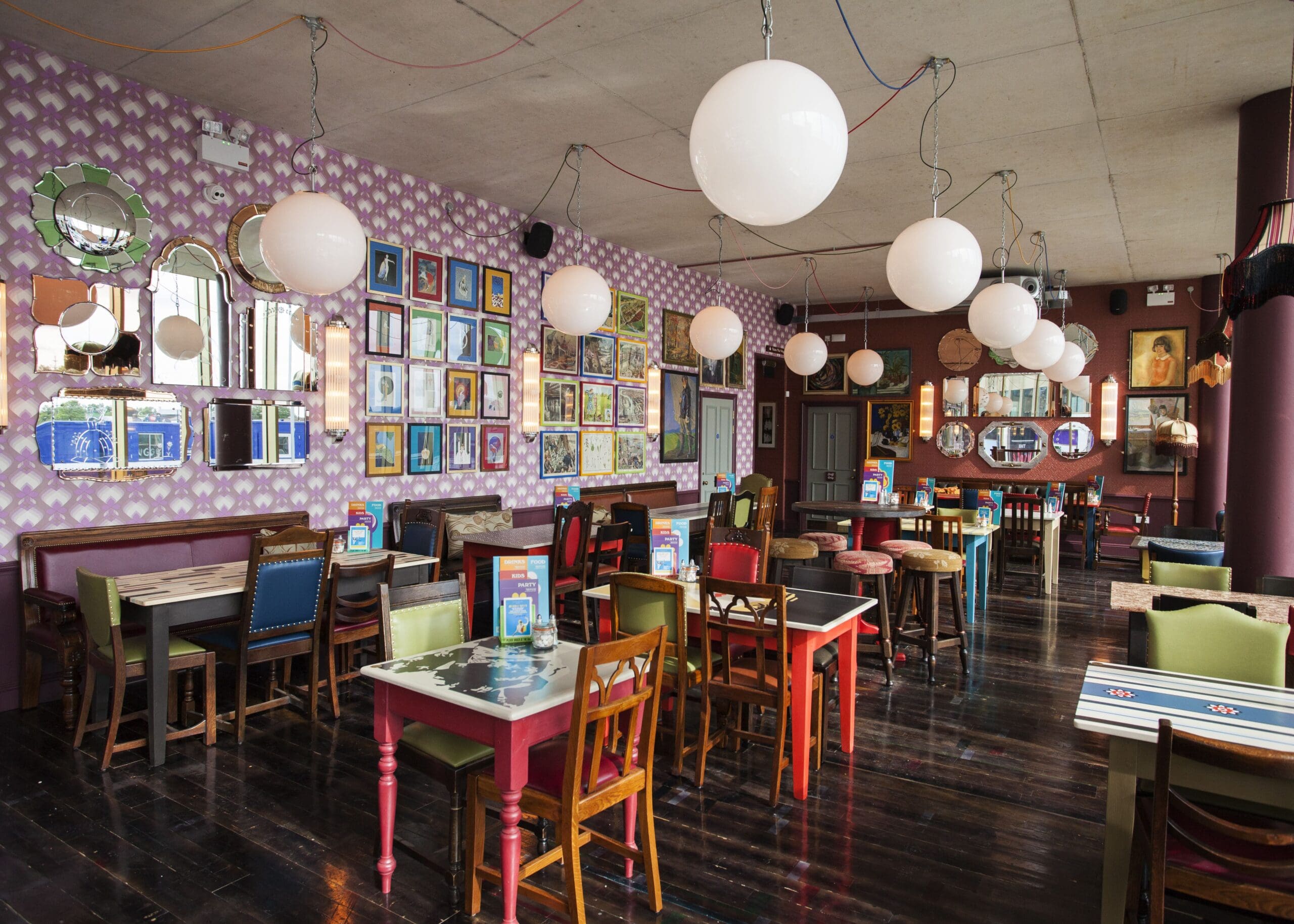 Colorful café interior with mismatched chairs and tables, round hanging lights, dark wooden floors, and walls decorated with many framed pictures and mirrors. The empty café exudes the unique charm of Falco style.