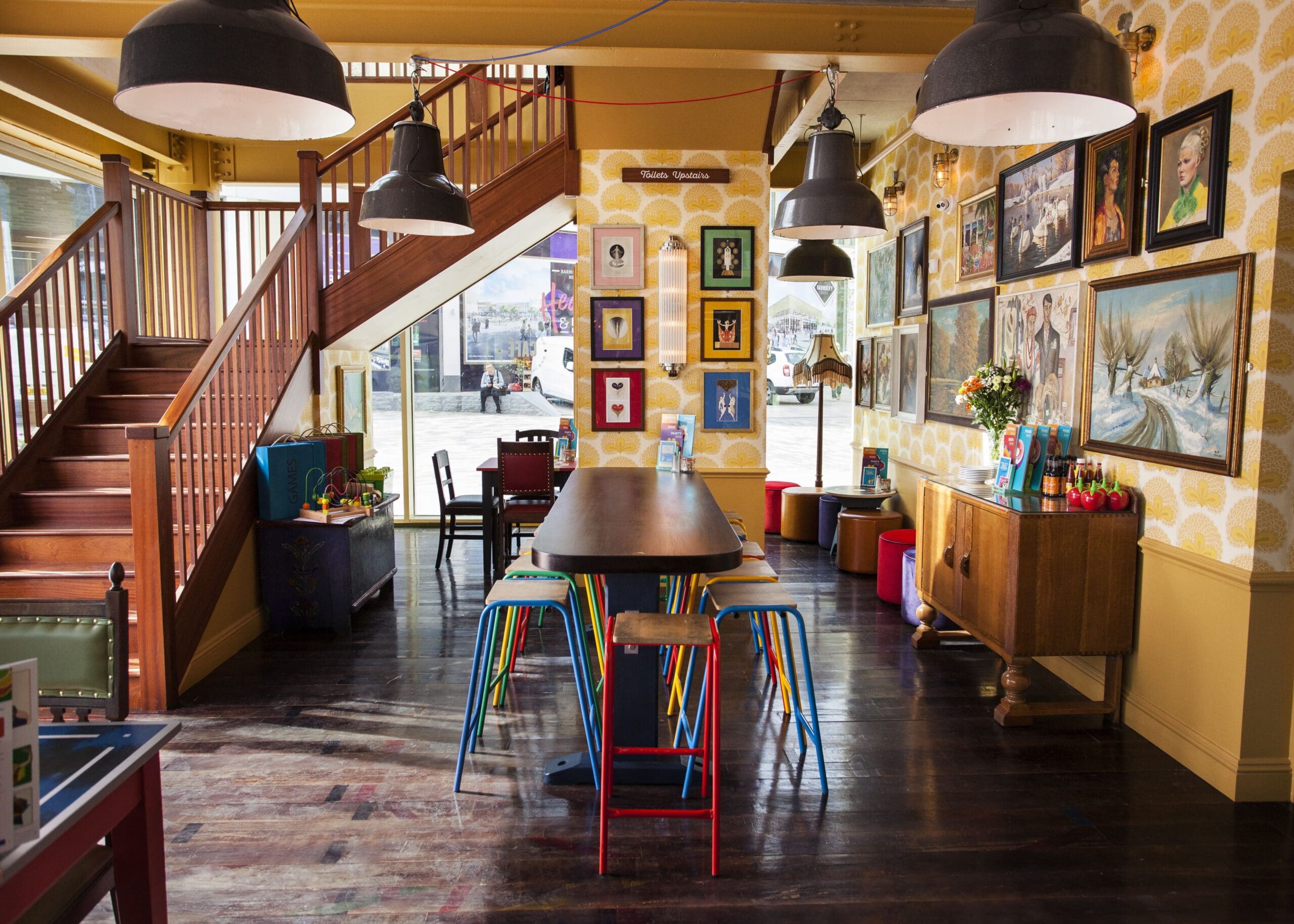 A cozy, colorful cafe interior with a long wooden table surrounded by multicolored stools, wooden stairs, Falco art prints on patterned walls, and large windows letting in natural light.