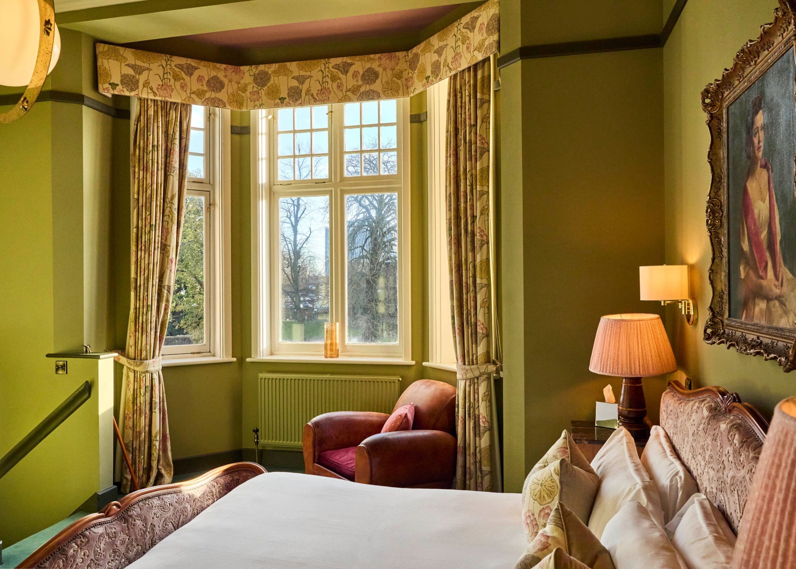 A cozy bedroom with green walls, a large bay window with curtains, an armchair, a bed with pillows, a bedside lamp, and a portrait painting on the wall. Sunlight streams in, creating a warm and inviting atmosphere.