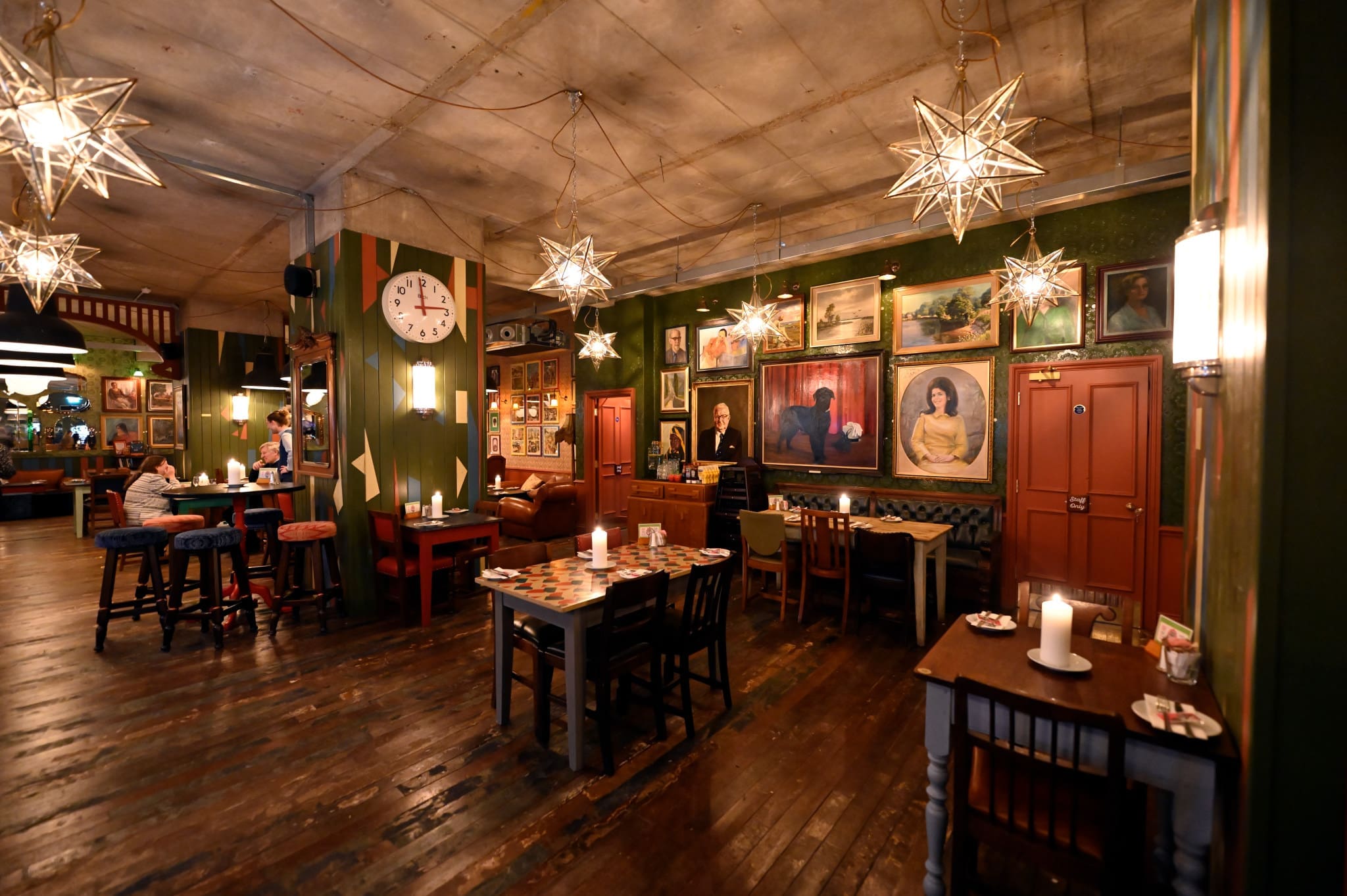 A cozy, dimly lit bar with wooden floors, eclectic wall art, Ferro accents, hanging star-shaped lights, and candles on the tables. Several tables and chairs are arranged around the room, creating a warm, inviting atmosphere.