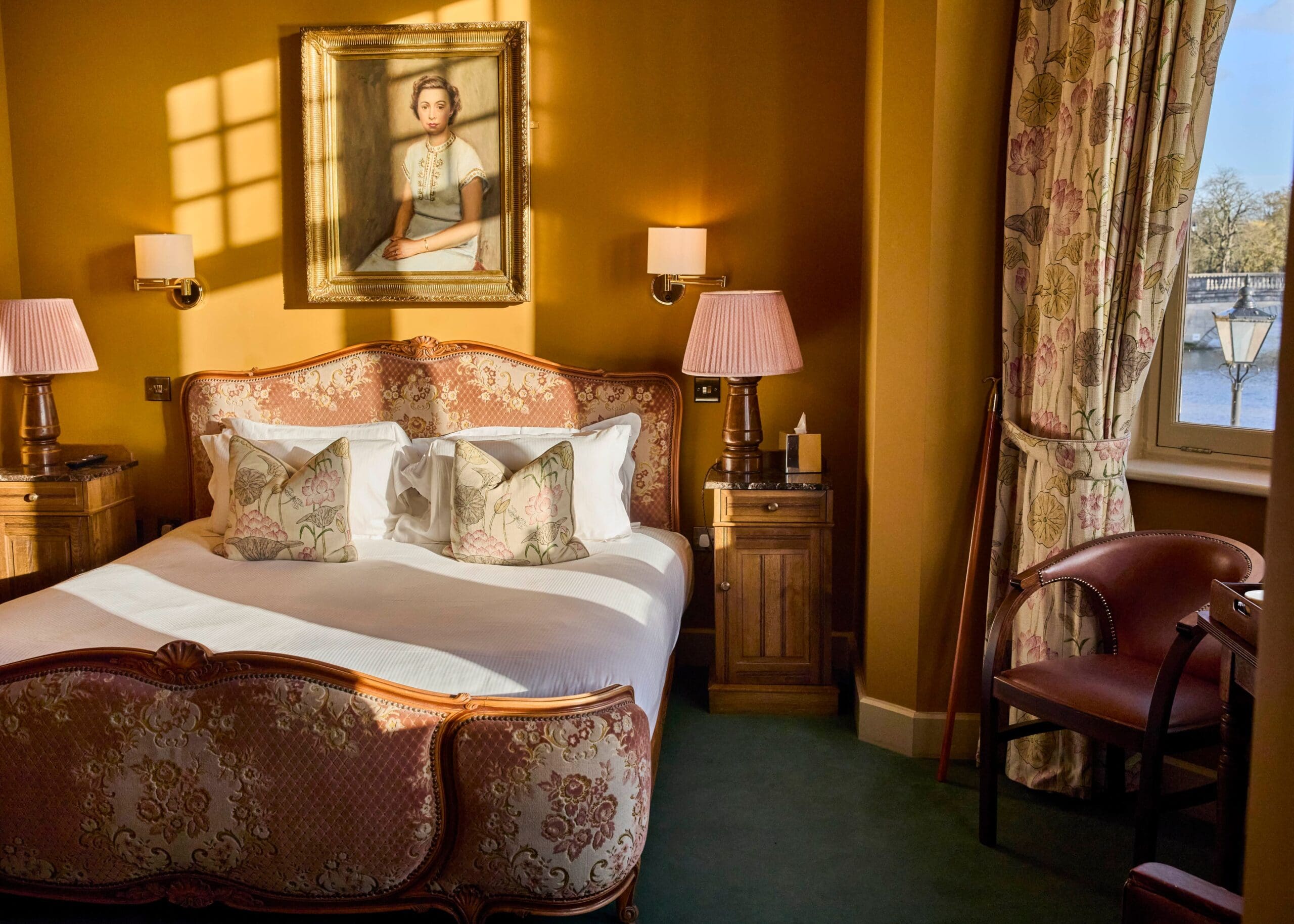 A warmly lit bedroom with a vintage-style bed, floral cushions, two wooden nightstands with lamps, a floral curtain, a desk with a chair, and a portrait of a woman hanging above the bed. Sunlight streams in through a window.