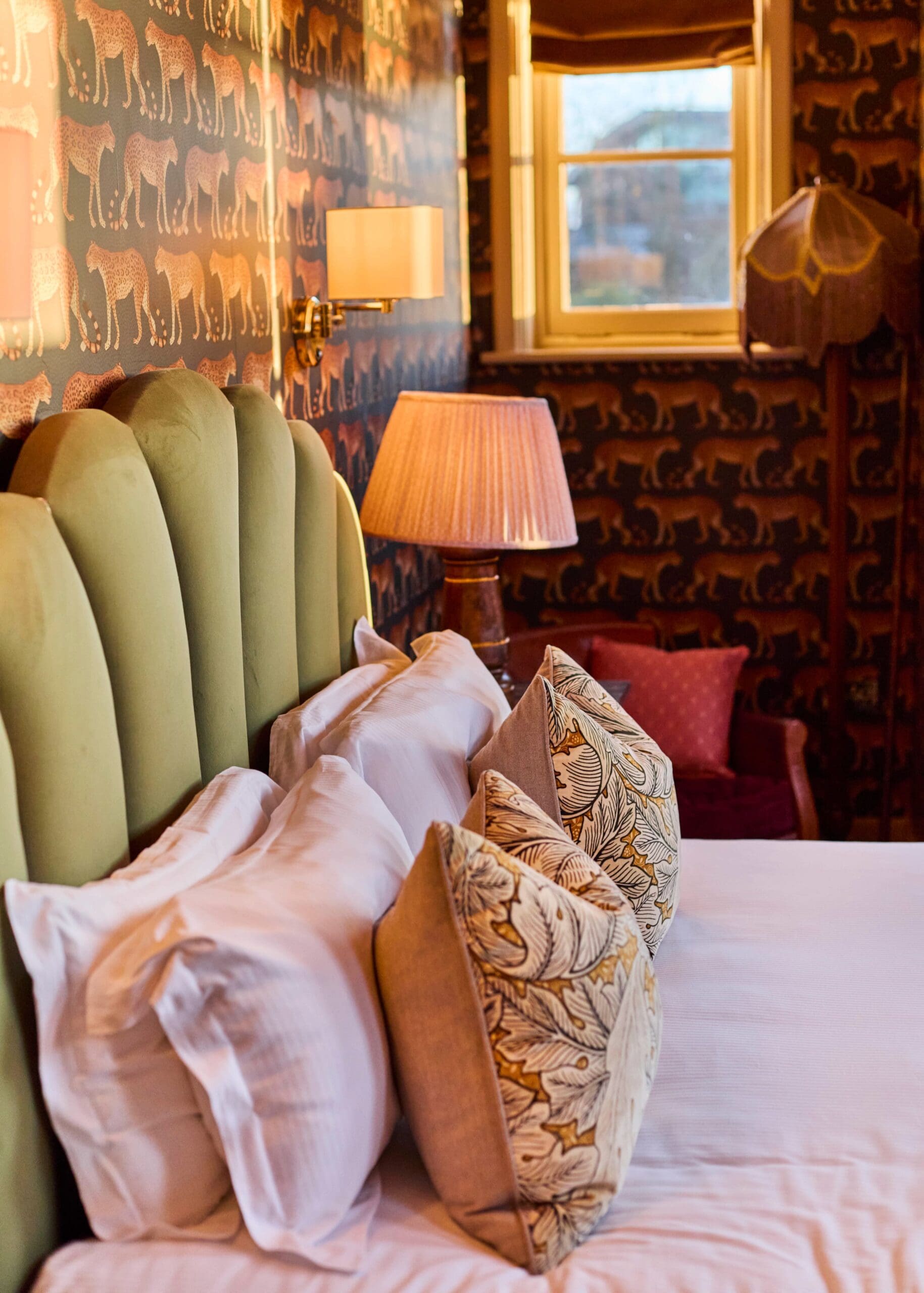 A cozy bedroom with a green upholstered headboard, patterned pillows, and a bed covered with white linens. The wallpaper features a repeating animal motif, and the room is softly lit by lamps, with a window in the background.