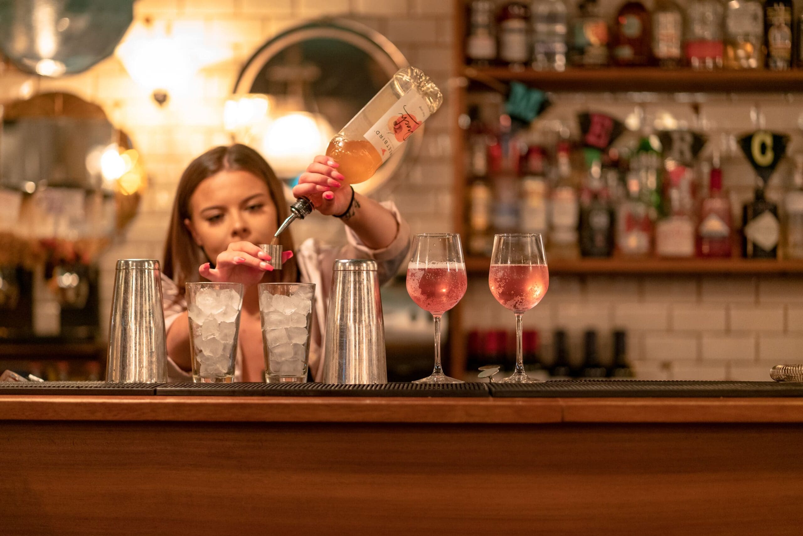 A bartender explores careers in mixology as they pour a bottle of rosé wine into a metal shaker filled with ice at a bar, with two glasses of rosé wine and bottles on shelves in the background.
