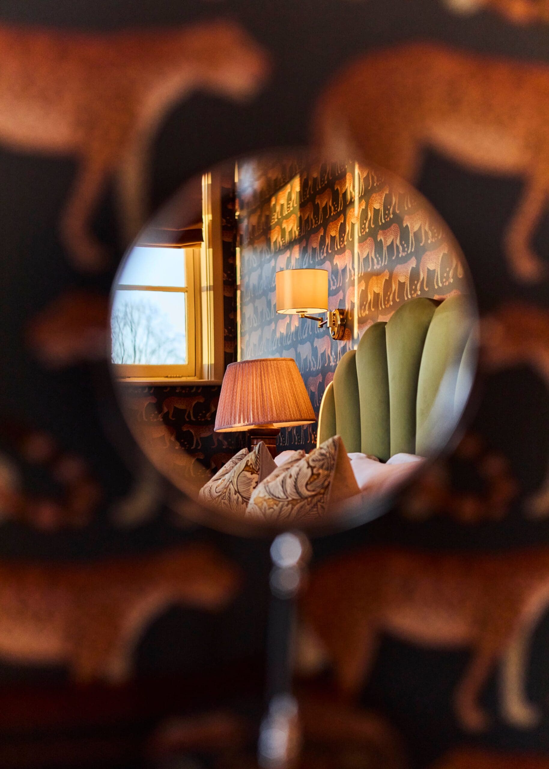A round mirror reflects part of a cozy Torino bedroom with a green upholstered headboard, patterned pillows, a lamp with a brown shade, and wallpaper featuring leopard designs.