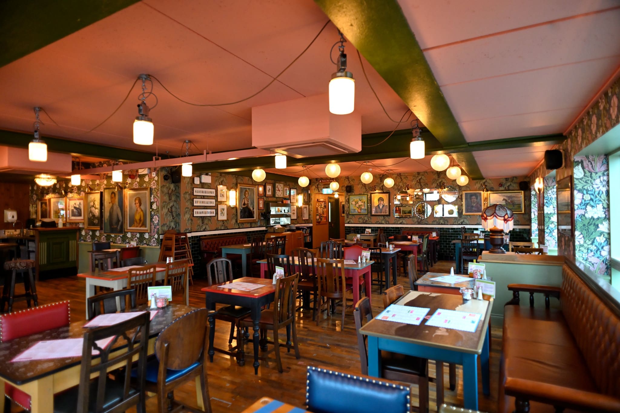 A cozy, eclectic restaurant interior, Fioro features colorful chairs, wooden tables, hanging lights, and framed artwork on patterned walls—creating a warm, inviting atmosphere. The space appears empty, with no people present.