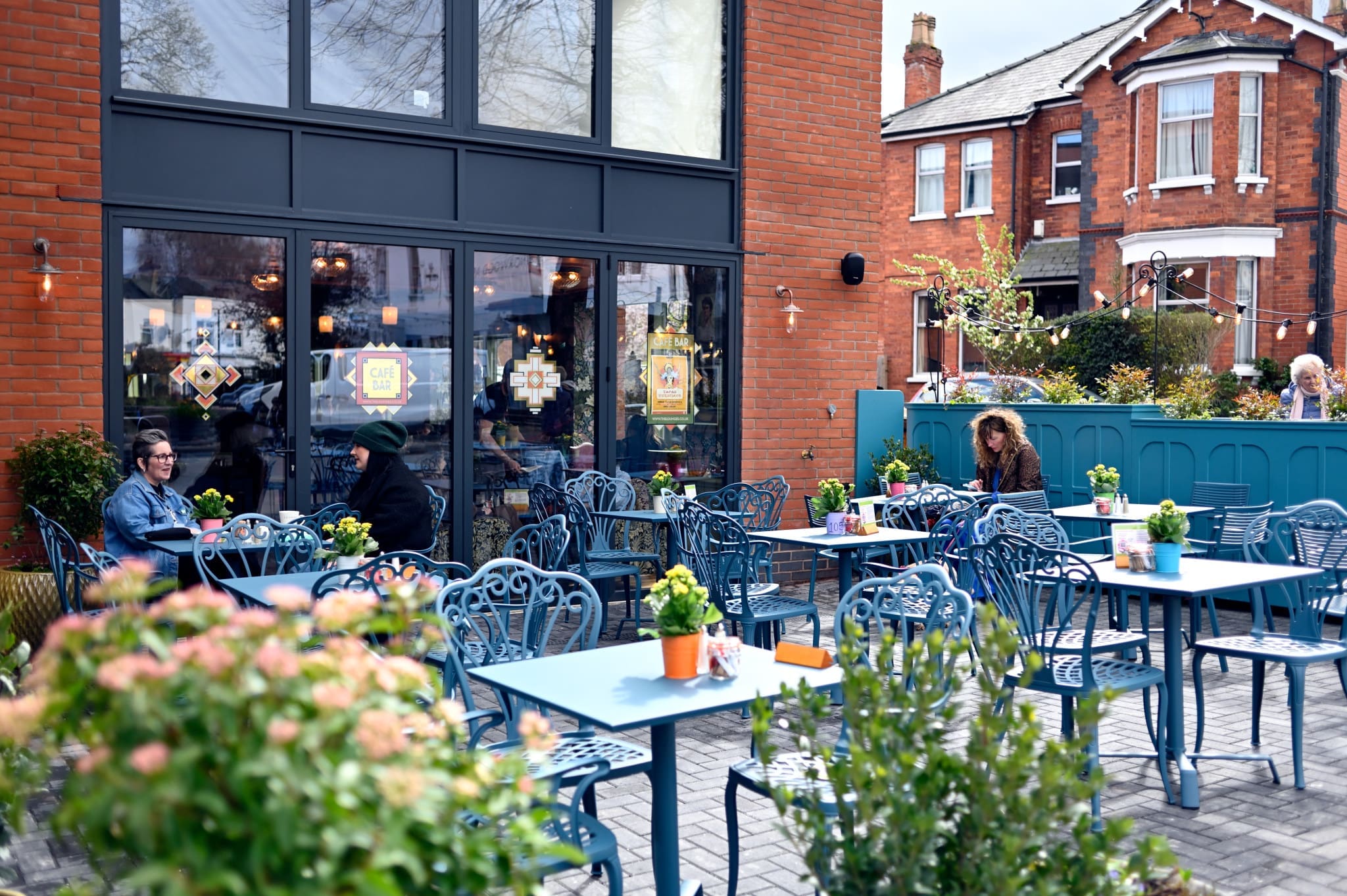 People sit at blue metal tables in the Fioro outdoor café, each table adorned with potted plants, beside a brick building. Residential houses and greenery are visible in the background on a sunny day.