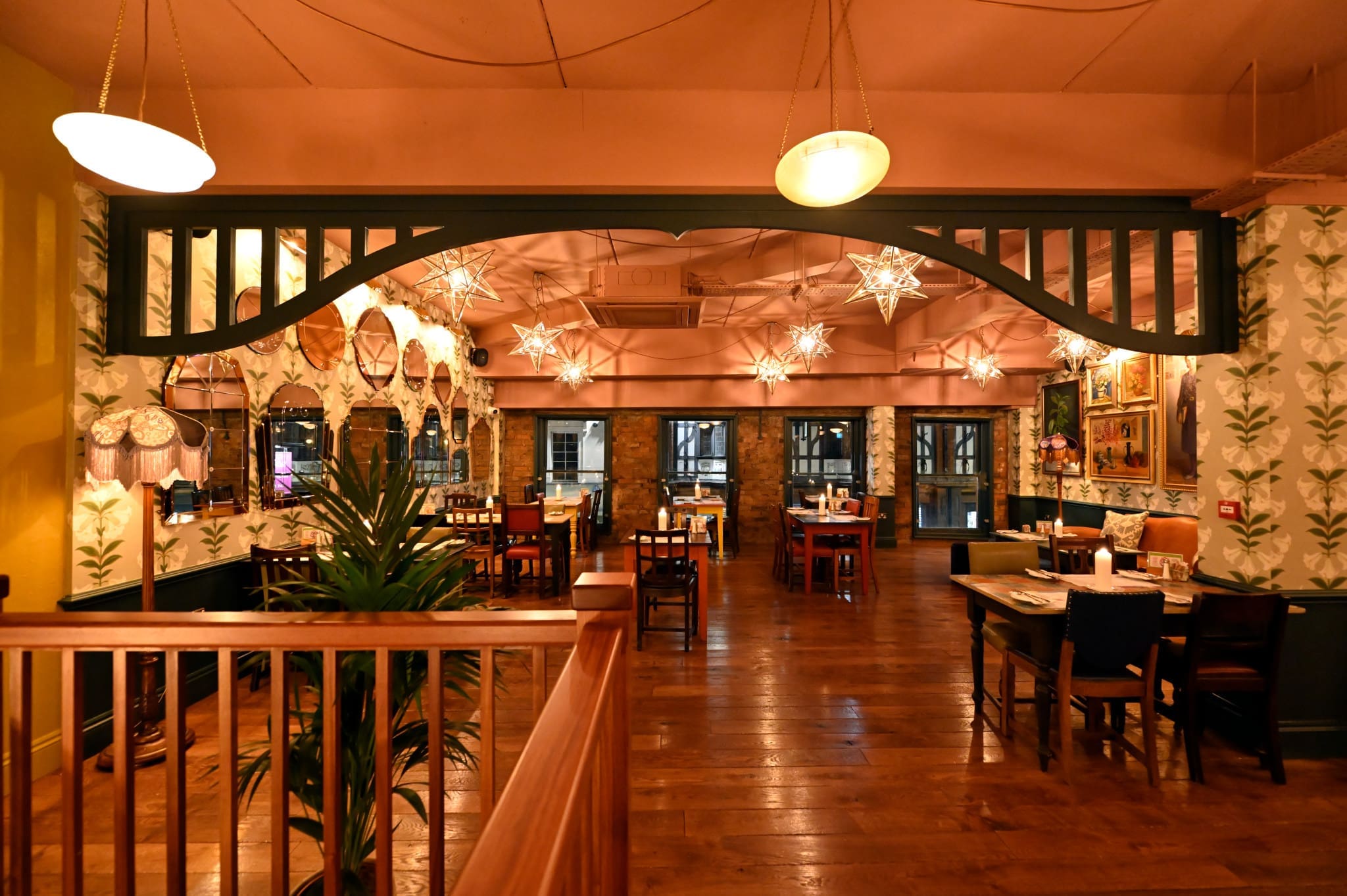 A warmly lit Floro restaurant interior with wooden floors, patterned wallpaper, star-shaped hanging lights, and neatly arranged tables and chairs, creating a cozy and inviting atmosphere.