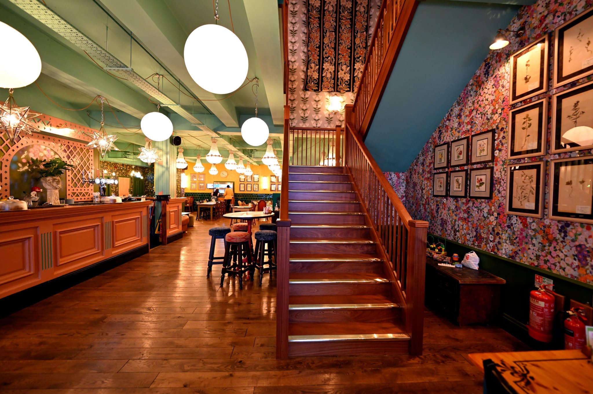 A warmly lit, vintage-style interior features Floro wooden floors, a staircase with floral wallpaper, framed art on the walls, and round hanging lights above tables and chairs in a cozy, inviting restaurant or café setting.