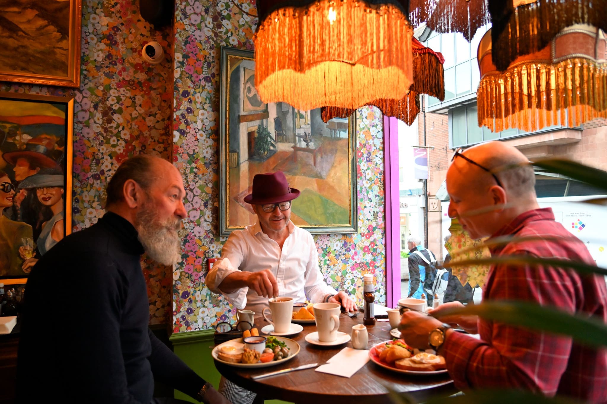 Three men sit around a small table in Floro, a cozy, colorful cafe with floral wallpaper, art, and warm hanging lamps, enjoying breakfast and coffee together. One man wears a purple hat and glasses.