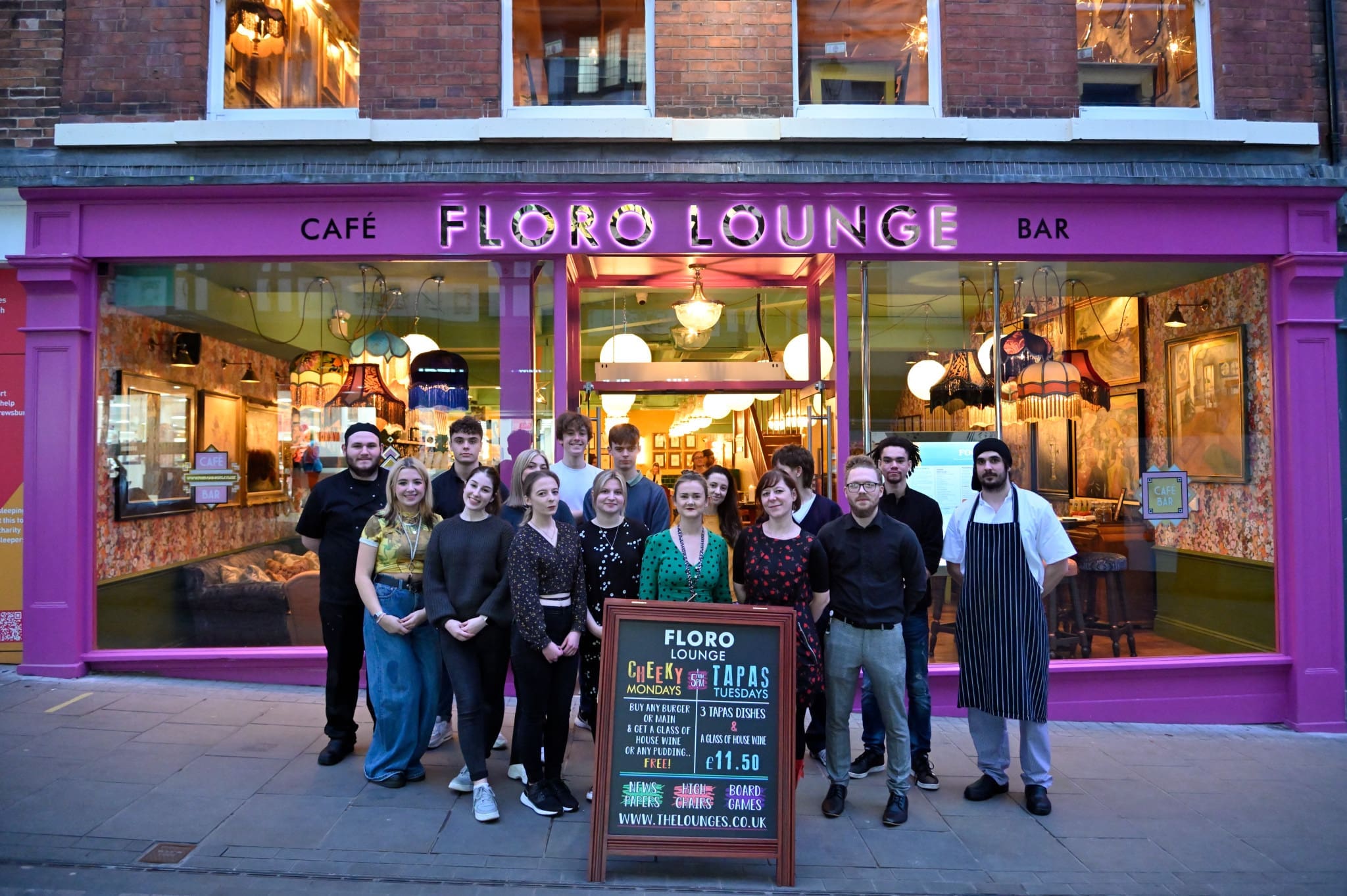 A group of people stands smiling in front of the vibrant, well-lit entrance of Floro Lounge Café Bar, with a chalkboard sign featuring Floro’s menu specials inviting guests from the sidewalk.