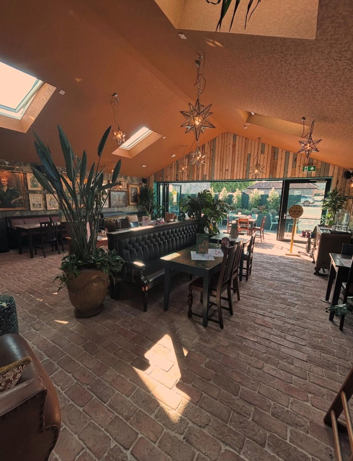 A cozy café with brick flooring, plant decorations, star-shaped hanging lights, and a mix of sofas and wooden chairs. Sunlight streams in through skylights and large windows, creating a warm, inviting atmosphere.