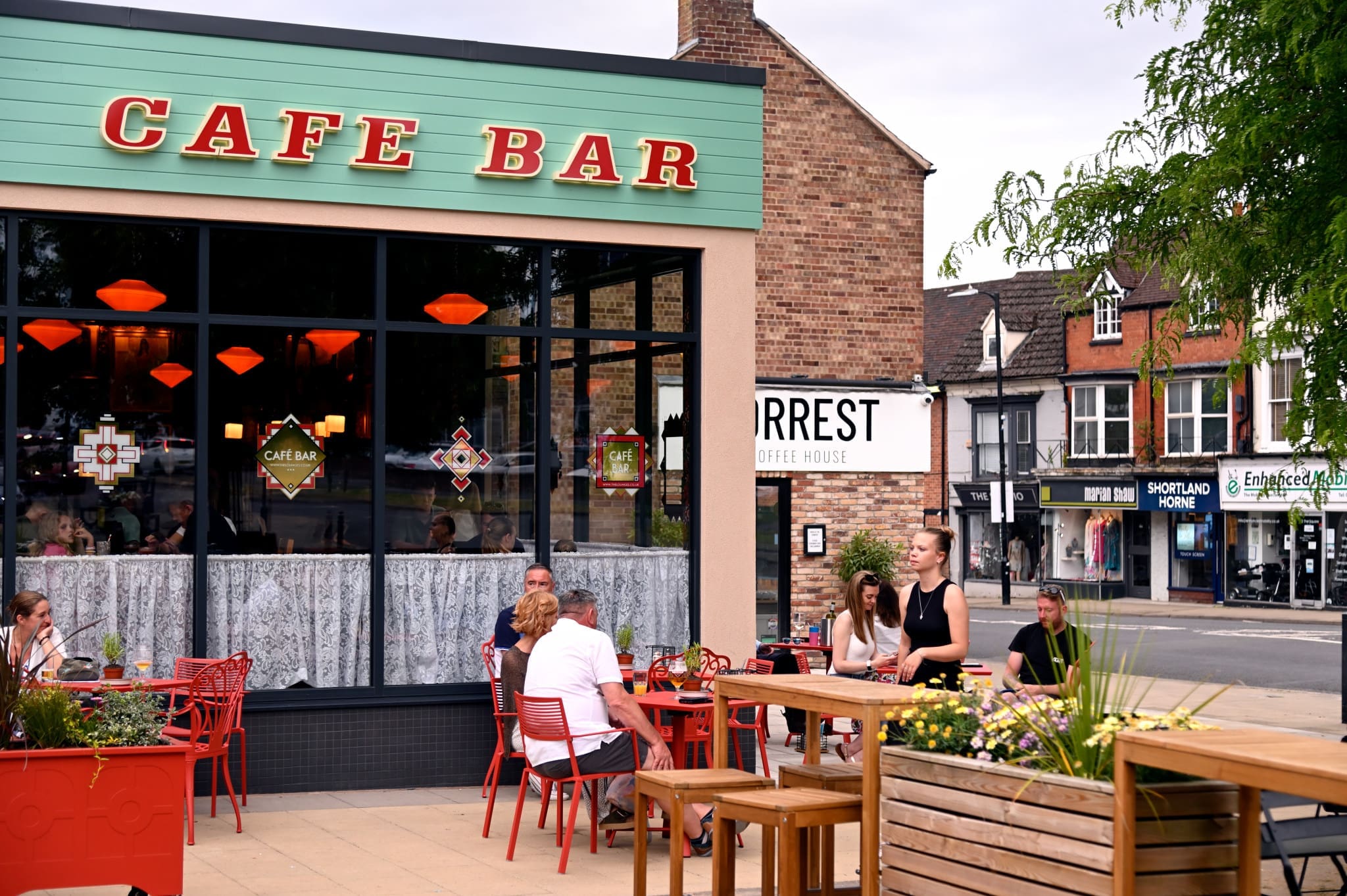 People sit at red tables outside a café bar on a sunny day in Fossato. A server stands near a wooden table with plants. The cafe has large windows, and a street with shops and houses is visible in the background.